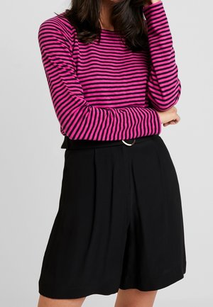 Kobieta ubrana w różowo-czarną bluzkę w paski z długim rękawem oraz czarne szorty z wysokim stanem z ozdobnym paskiem z przodu.