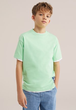 Chłopiec o jasnobrązowych włosach, ubrany w jasnozieloną koszulkę na białej koszuli i niebieskich dżinsach, stojący na jednolitym beżowym tle.