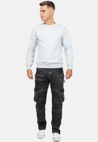 Felpa grigia con polsini a costine, abbinata a pantaloni cargo neri con molteplici tasche con zip e dettagli testurizzati. Indossato con sneakers bianche.