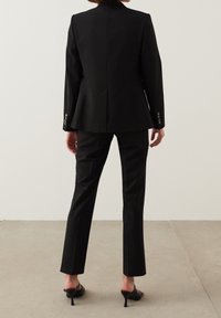 Blazer noir ajusté et pantalon droit. Le costume présente un design épuré, une texture lisse et des accents de boutons argentés subtils.