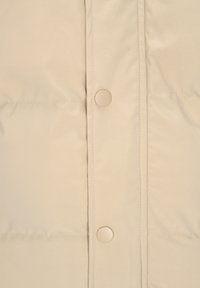 Beige polstret jakke med glat, mat stof. Har runde, lyse knapper og syede sektioner for ekstra tekstur.