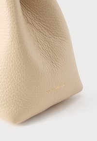 Γωνία μπεζ τσάντας από υφή δέρματος με διακριτικό χρυσό λογότυπο "COCCINELLE" πάνω σε λευκό φόντο.