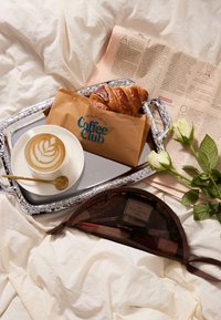 Un vassoio argentato contiene un latte in una tazza bianca con un motivo a foglia, un croissant, una busta di carta marrone e un giornale rosa con delle rose accanto.