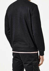 Homme portant un sweat-shirt noir et un pantalon cargo noir, dos tourné, sur un fond blanc uni.