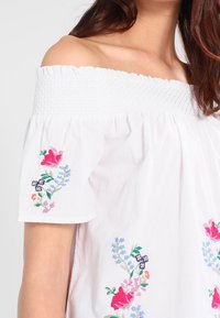 Donna che indossa una blusa bianca senza spalline con colletto arricciato e ricami floreali colorati sulle maniche e sulla parte anteriore.