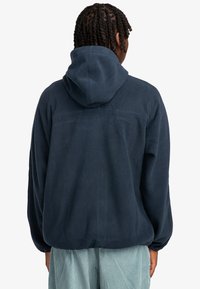 Mørkeblå fleece hoodie med hætte, løs pasform og tekstureret stof. Har en søm over den øverste del af ryggen og ribbestrikkede manchetter.