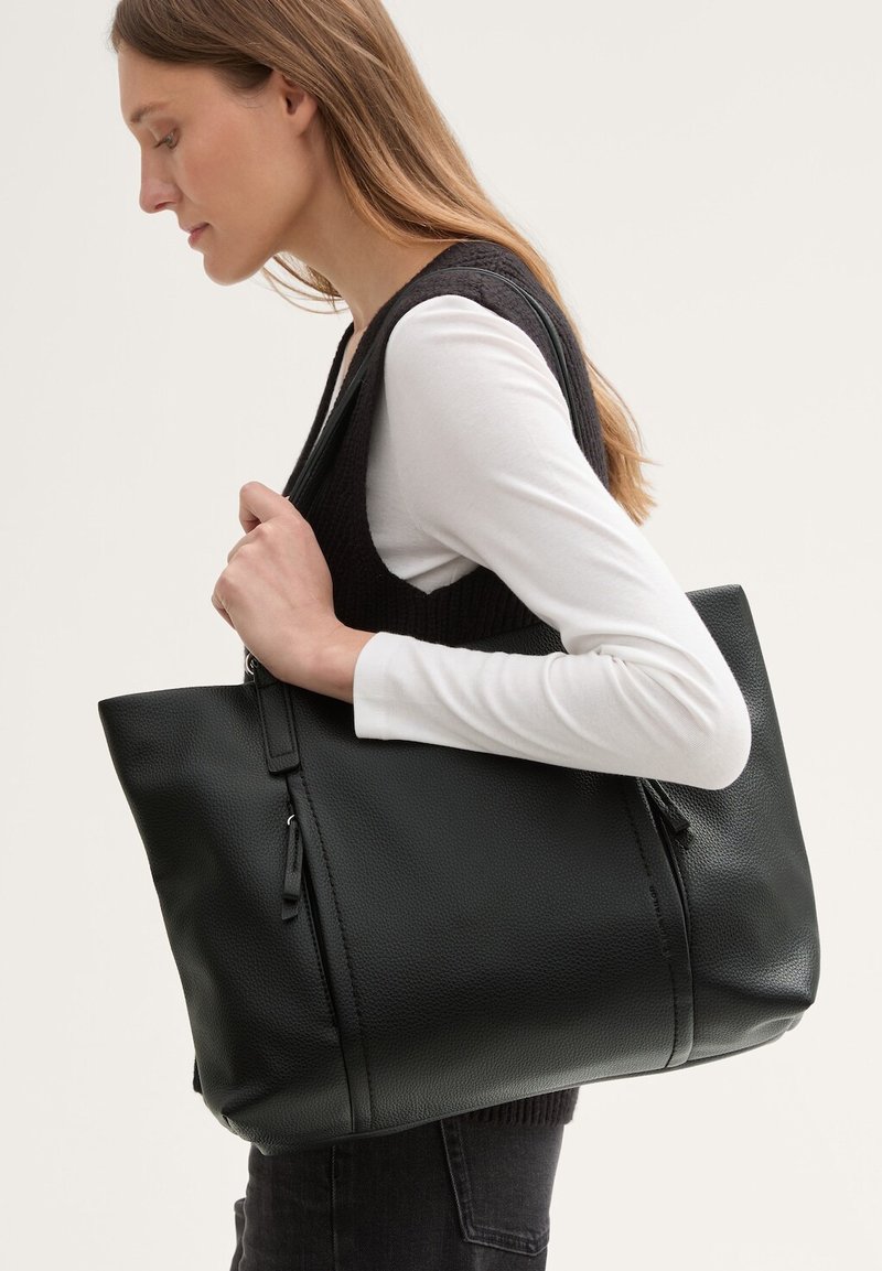 Zwarte leren tote bag met een gestructureerde afwerking, grote vorm, ritsdetails en dubbele handvatten aan de bovenkant, vastgehouden door een persoon in profiel.