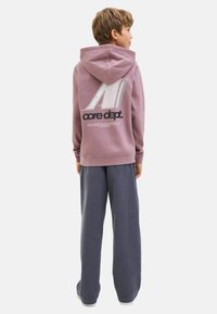 Un enfant aux cheveux ébouriffés se tient de dos, portant un hoodie rose avec un motif graphique et un pantalon large gris.