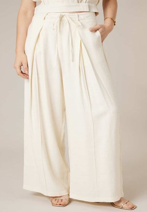 Femme portant un pantalon crème taille haute à jambes larges avec des plis à l'avant et une ceinture en tissu nouée à la taille, associée à des sandales dorées à lanières.