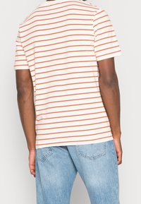 Hombre con camiseta blanca de rayas horizontales rojas finas y jeans de denim azul claro, visto desde atrás contra un fondo sencillo.