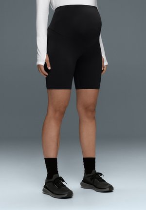 Personne enceinte portant un short de vélo de maternité noir, des chaussettes noires, des chaussures de course noires et un haut blanc à manches longues sur un fond gris.