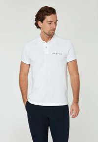 Weißes Polo-Shirt aus Baumwolle mit Kragen, zwei Knöpfen und einem kleinen Logo auf der linken Brust. Getragen mit dunkelblauen Hosen.