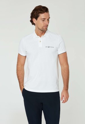 Weißes Polo-Shirt aus Baumwolle mit Kragen, zwei Knöpfen und einem kleinen Logo auf der linken Brust. Getragen mit dunkelblauen Hosen.