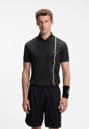 Muškarac nosi crnu polo majicu sa tankim okomitim prugama i tekstom "BOSS", crne kratke hlače i crnu narukvicu s tekstom "BOSS" na lijevom zapešću.