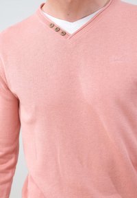 Pull en tricot rose clair avec un col en V, arborant trois boutons décoratifs à gauche et un logo subtil brodé sur le devant.
