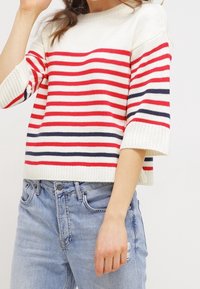 Suéter a rayas en crema, rojo y azul marino, con un cuello redondo y mangas cortas y anchas. Combinado con jeans desgastados de color azul claro.