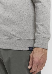 Grauer Sweatshirt mit strukturierter Oberfläche, ausgestattet mit einem gerippten Saum und Bündchen, und einem kleinen blau-weißen-roten Etikett an der Seite. Kombinierbar mit grünen Hosen.