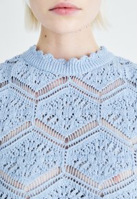 Ljusblått stickat linne med en vågig halsringning och hexagonalt hålmönster, som framhäver en mjuk textur och intrikata detaljer.