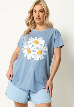 Lyshåret kvinde iført en løs blå t-shirt med tre hvide tusindfryd med gule centre kombineret med lyse denimshorts.