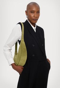 Bolso de ante verde oliva con un diseño curvado, llevado sobre un conjunto negro entallado. Presenta una textura suave y un minimalismo en los acentos de costura.