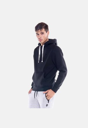 Jeune homme portant un sweat à capuche noir avec des cordons blancs et un pantalon de jogging gris clair, debout les mains dans les poches sur un fond blanc.