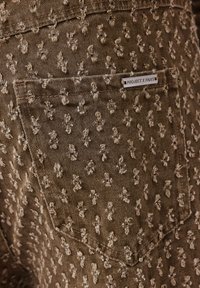 Tessuto di denim marrone con un motivo testurizzato di motivi in rilievo beige chiaro. Presenta una tasca etichettata con il tag "PROJECT X PARIS".