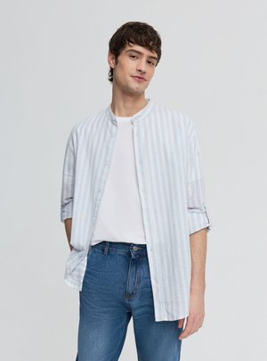 Młody mężczyzna w białej koszulce, jasnoniebiesko-białej paskowanej koszuli na guziki oraz niebieskich dżinsach, stojący z jedną ręką w kieszeni.