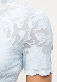 Διάφανο γαλάζιο ύφασμα με λουλουδένιο δαντελένιο μοτίβο που καλύπτει έναν κοντομάνικο ώμο και το πάνω μέρος του βραχίονα.