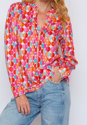 Femme portant une chemise colorée à manches longues boutonnée avec des motifs circulaires roses, orange et bleu sarcelle, et un jean bleu clair, main dans la poche.