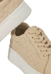 Baskets beiges avec texture tissée, bout rond et semelle en caoutchouc blanc. Comprend un système de laçage et des œillets dorés.