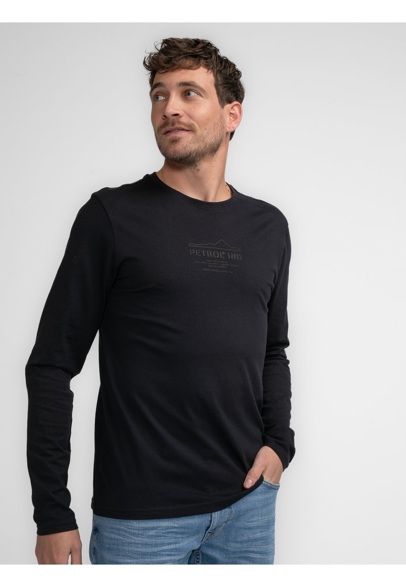 Tricou negru cu mânecă lungă, realizat dintr-un material moale, având un logo imprimat subtil pe piept, purtat cu blugi de denim albaștri deschiși.