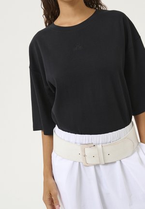 Vrouw draagt een zwart oversized T-shirt, ingestopt in witte broek met een brede witte riem met een grote vierkante gesp.