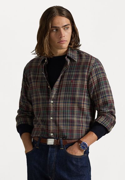 Chemise à carreaux pour homme dans des tons verts foncés, rouges et bleus, avec un col boutonné. Portée sur un haut noir, associée à un jean bleu et une ceinture brune.