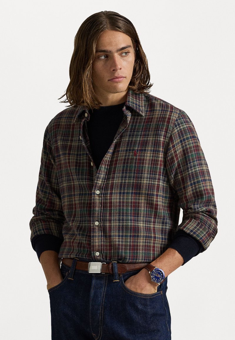 Heren geruit overhemd in donker groen, rood en blauwe tinten, met een button-down kraag. Draag het over een zwarte top, gecombineerd met blauwe jeans en een bruine riem.