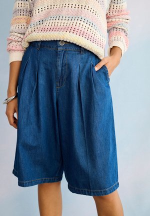 Osoba nosiaca modré plisované rifľové šortky a pastelovo pruhovaný pletený sveter s jednou rukou v vrecku na svetlom pozadí.