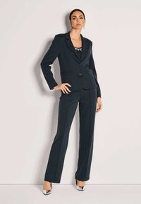 Marineblauer, maßgeschneiderter Anzug mit tailliertem Blazer und gerade geschnittenen Hosen, versehen mit dezenten kontrastierenden Nähten und silbernen Akzenten an den Schuhen.