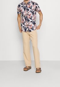 Korte mouwen, marineblauw bloesje met roze en witte patronen, gecombineerd met beige broek en bruine sandalen met verstelbare bandjes.