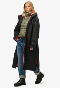 Abrigo largo negro con detalles rojos sobre una chaqueta acolchada verde, combinado con jeans azules arremangados y botas negras de cordones. Tejido suave, diseño en capas.