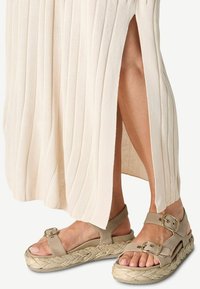 Piedi che indossano sandali con plateau beige con cinturini a fibbia, parzialmente coperti da una gonna a coste color crema con uno spacco laterale.