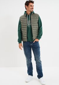 Gilet grigio imbottito sopra un felpa verde, jeans blu e sneakers bianche. Il gilet presenta cuciture orizzontali e un dettaglio a logo rotondo.
