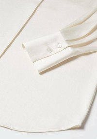Cream-colored long-sleeve shirt with a buttoned cuff featuring two white buttons, crafted from smooth fabric with a subtle texture.