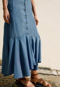 Denim maxi kjole med knaplukning foran, flared kant og flæset bund. Bæres med brune sandle med stropper, der har en tyk sål og ankelstropper.