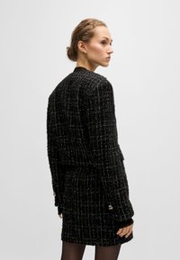 Musta tweed-takki, jossa on tekstuuri kuvio, kultaiset yksityiskohdat hihoissa, yhdistettynä vastaavaan tweed-hameeseen.