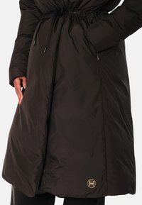 Svart polstret kåpe med snor i livet, lange ermer og sidelommer. Har en gullfarget emblemdetalj nær kanten. Glatt tekstur.