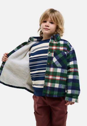 Enfant blond portant un pull à rayures bleues et blanches et une veste à carreaux verts, tenant la veste ouverte pour montrer sa doublure en polaire blanche.