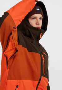 Waterdichte jas in bruin en oranje, met een ritssluiting aan de voorkant, een verstelbare capuchon en mesh gevoerde zakken voor ventilatie.