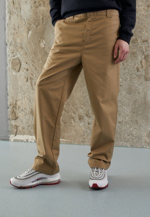 Beige chinos med lige pasform, lavet af glat stof, med sidelommer og iført grå og røde sneakers.