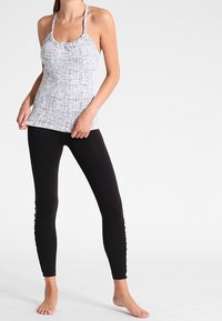 Světlý šedý tílko s texturovaným vzorem a tenkými ramínky, kombinovaný s černými legínami s krajkovým šněrováním podél bočních švů.