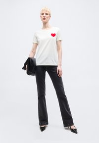 Ξανθός άνθρωπος που στέκεται, φοράει λευκό μπλουζάκι με λεπτομέρεια κόκκινης καρδιάς, μαύρα τζιν, μαύρες μυτερές γόβες και κρατάει διπλωμένο μαύρο σακάκι στο ένα χέρι.