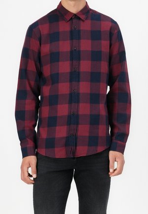 Uomo che indossa una camicia a maniche lunghe con un grande motivo a quadri rosso e nero e pantaloni neri su uno sfondo uniforme.